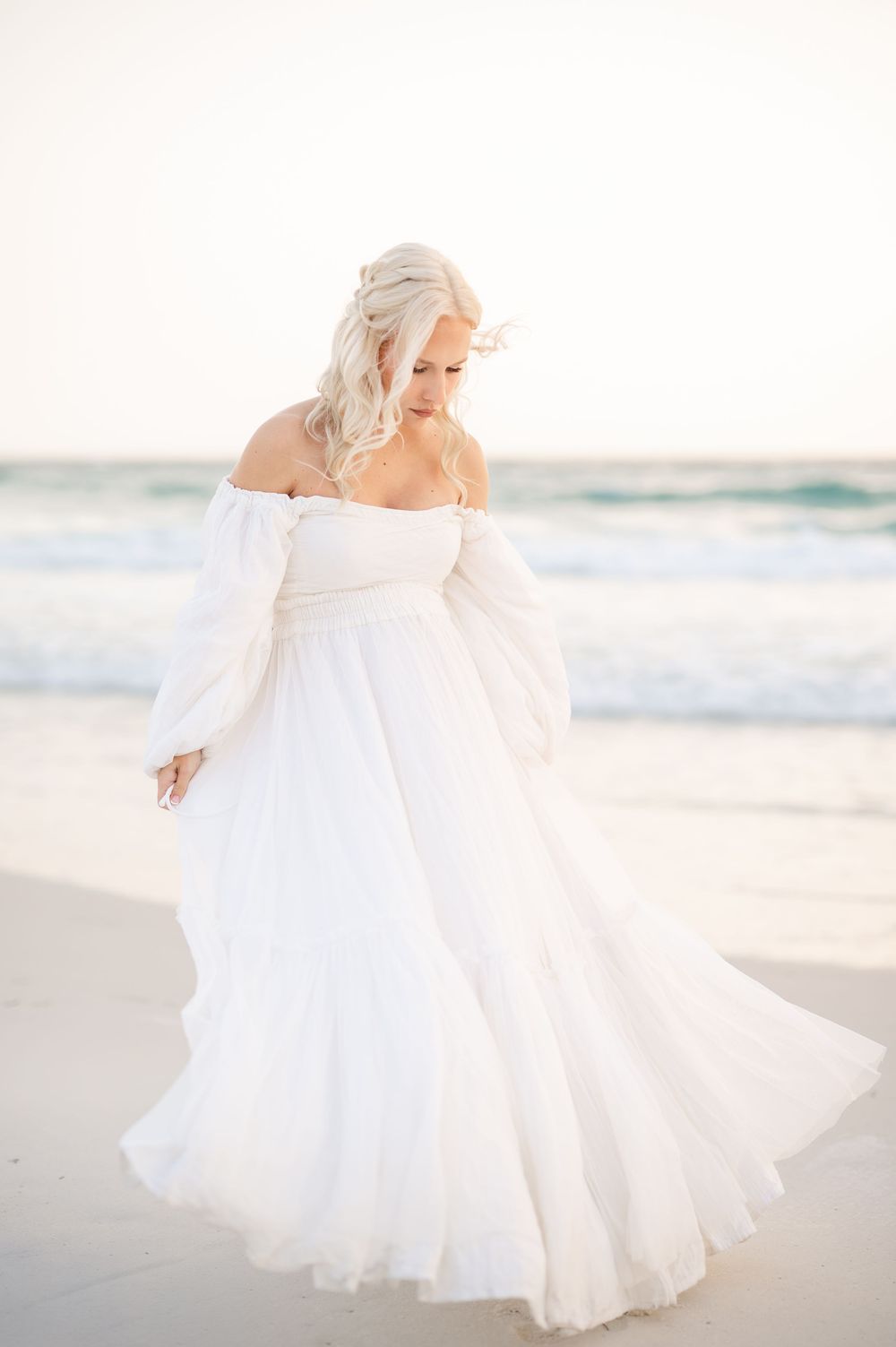 Bride in flowing white off-shoulder gown twirling on a sandy beach with ocean waves in the background.