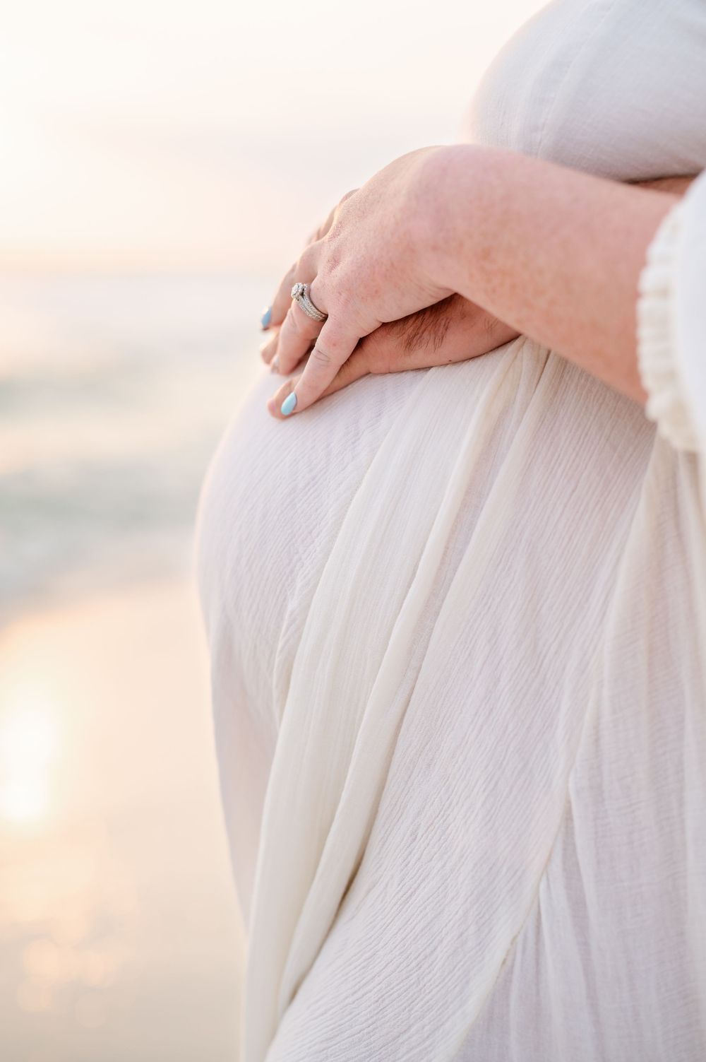 Close-up of a pregnant woman in a white dress cradling her baby bump on a serene beach at sunset.