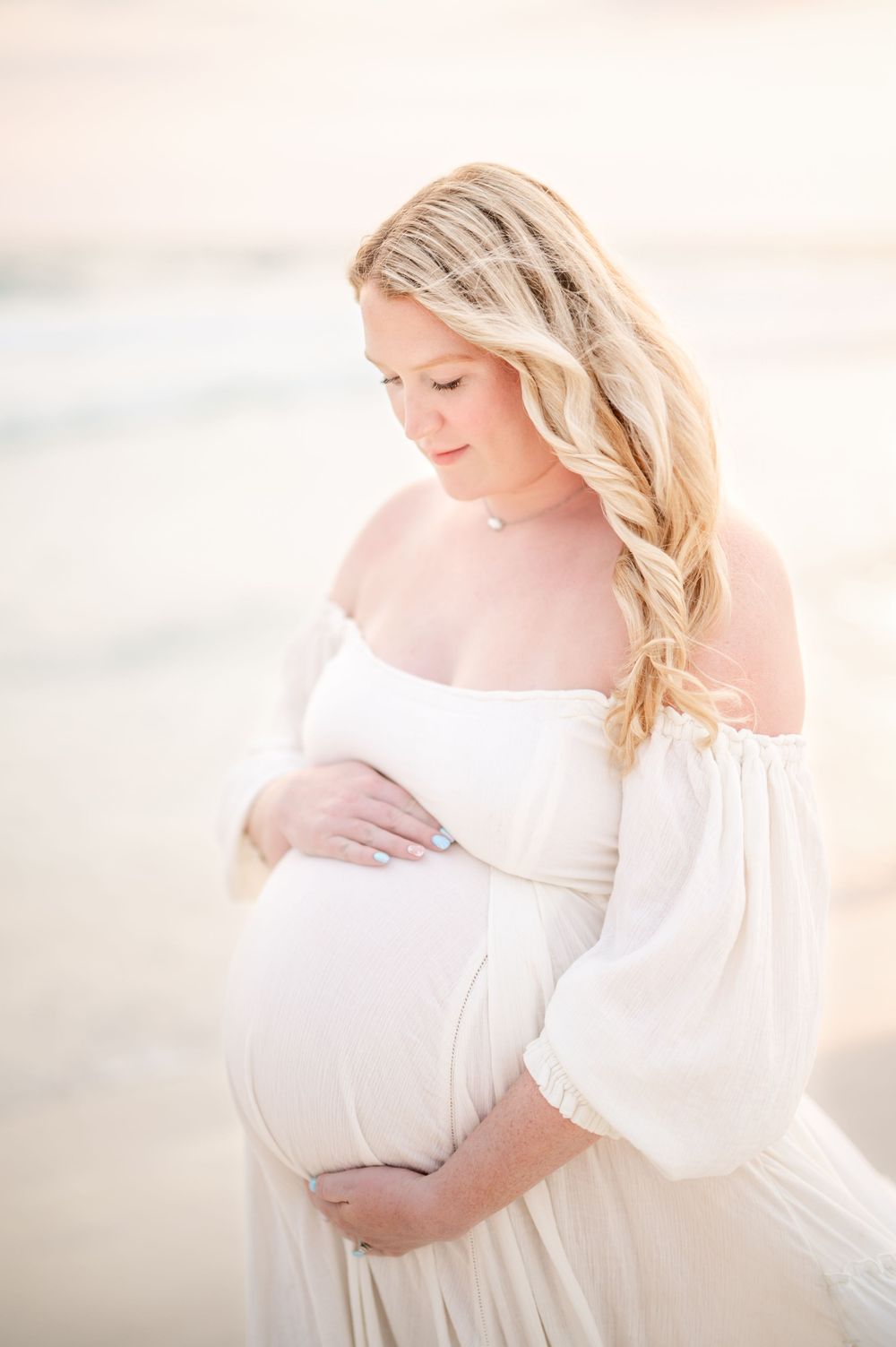 Pregnant woman in white off-shoulder gown cradling baby bump on beach at golden hour.