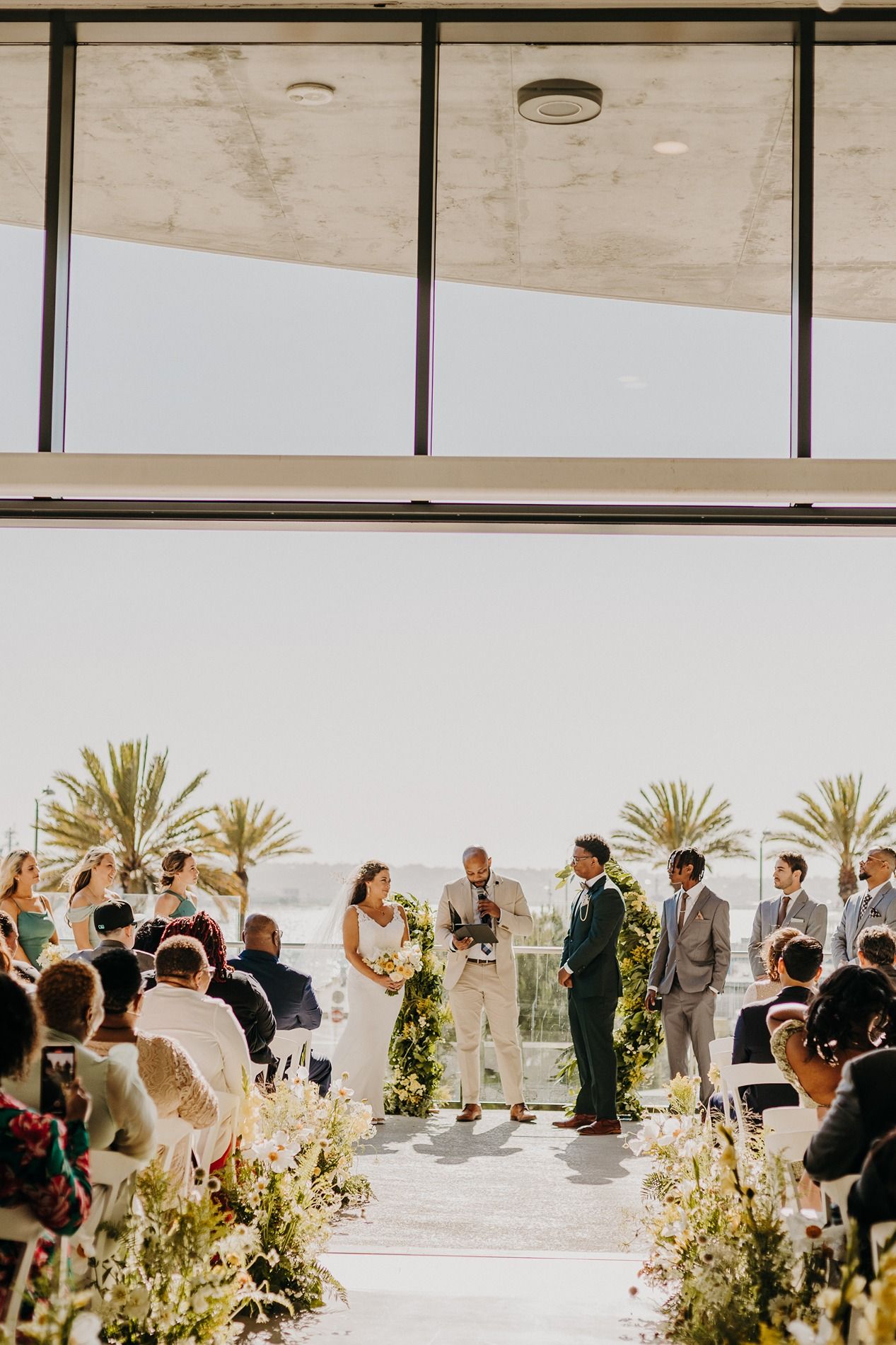 Outdoor wedding ceremony with couple at altar, lush greenery, palm trees, and ocean view backdrop.