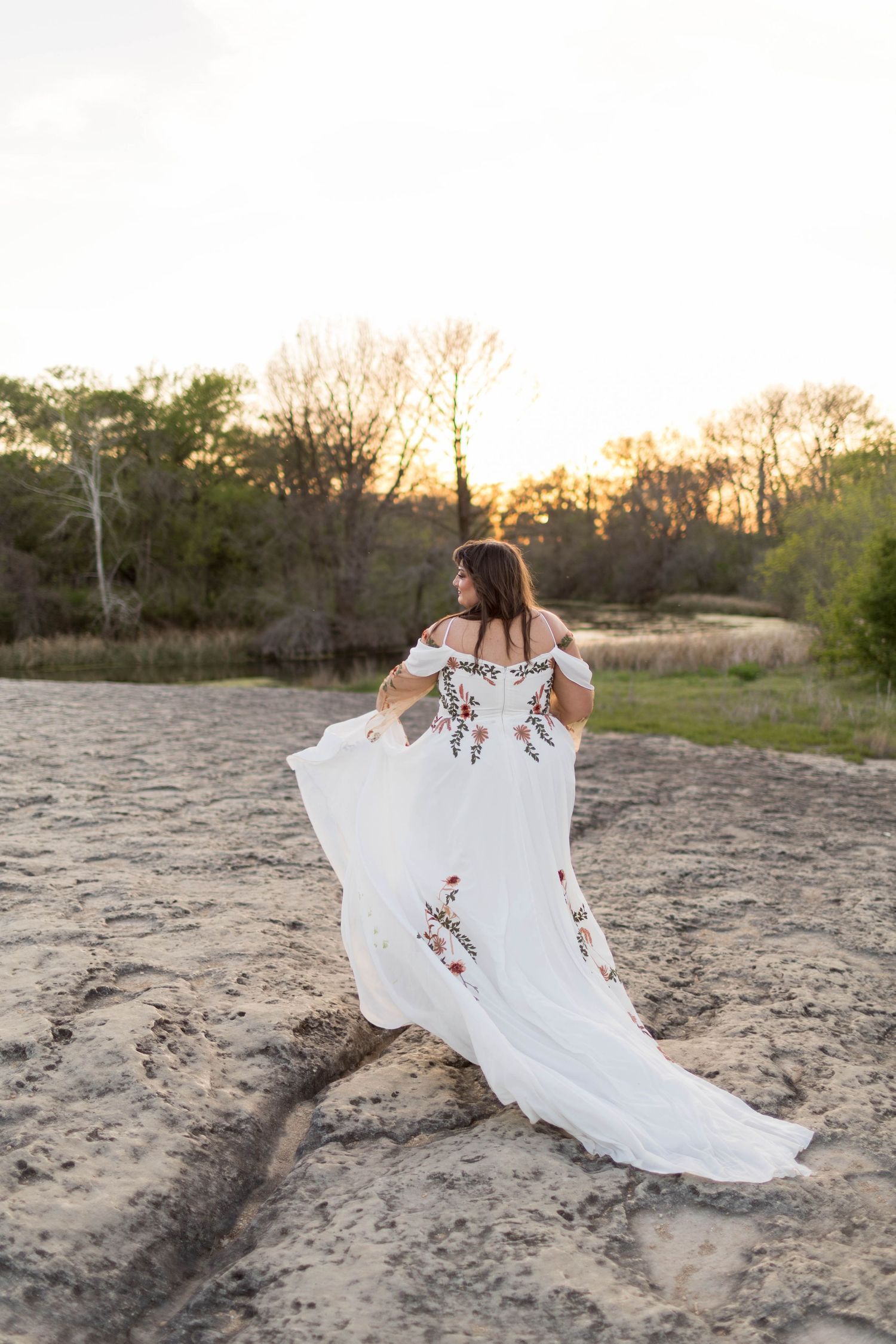 Woman in white floral embroidered off-shoulder gown poses on sandy terrain at golden hour with trees in background.