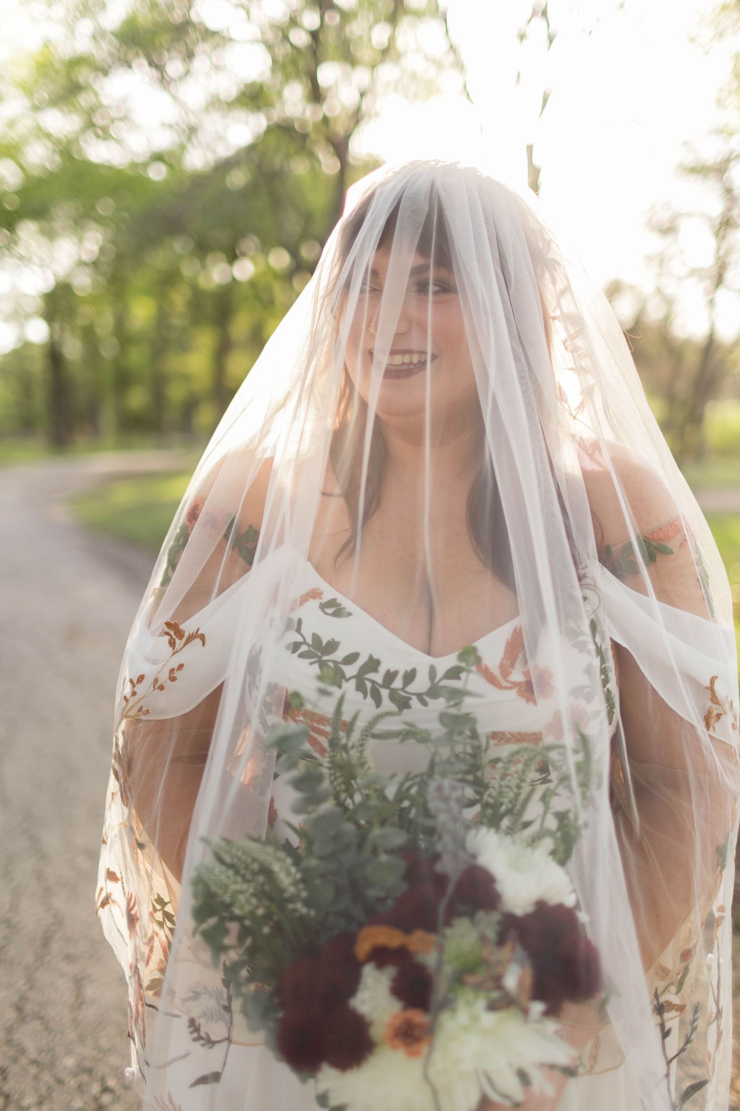 Smiling bride in floral wedding dress and veil holds bouquet of burgundy and white flowers outdoors.