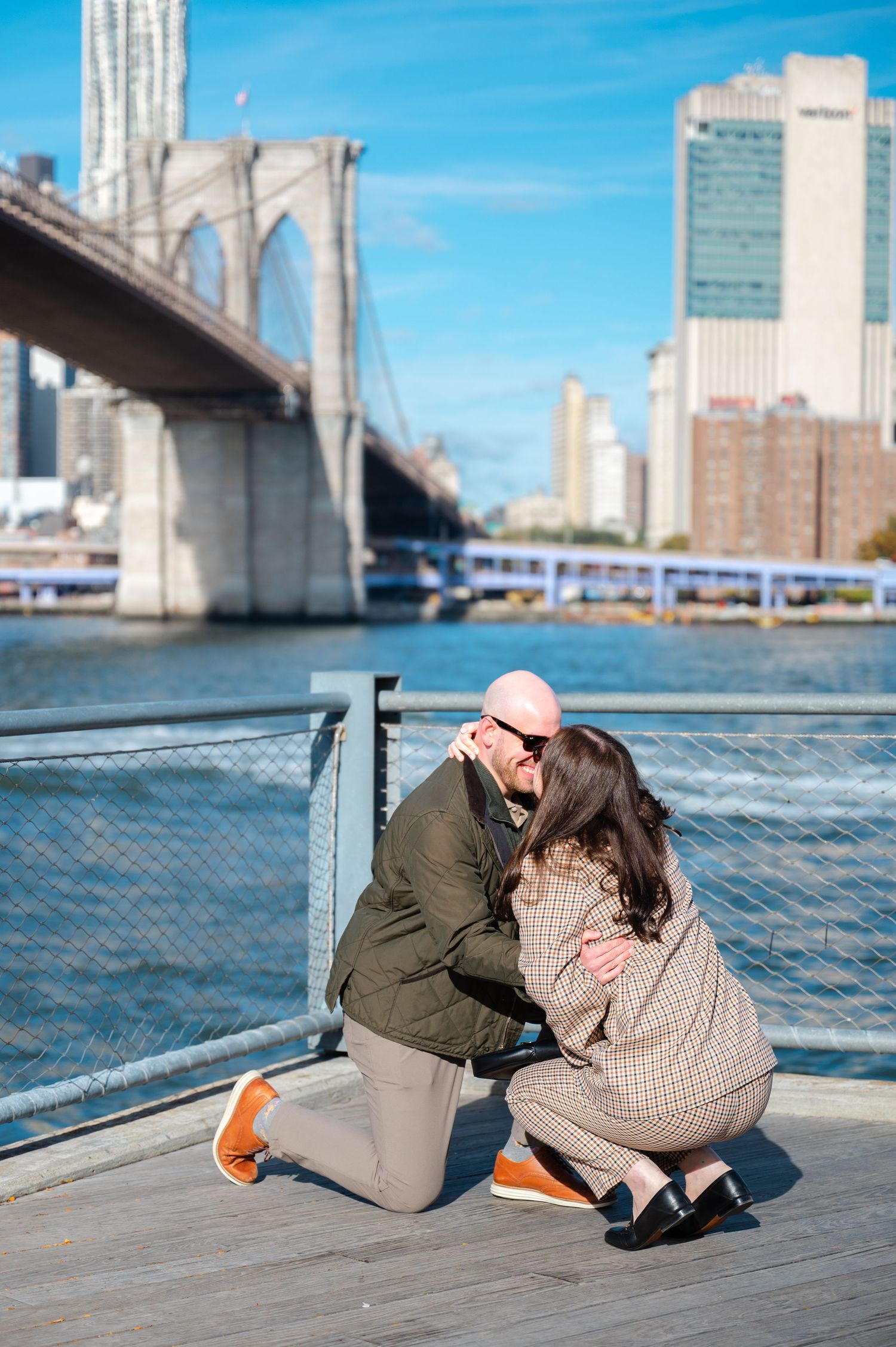 Couple sharing a romantic proposal moment on a wooden pier with the Brooklyn Bridge and NYC skyline in the background.