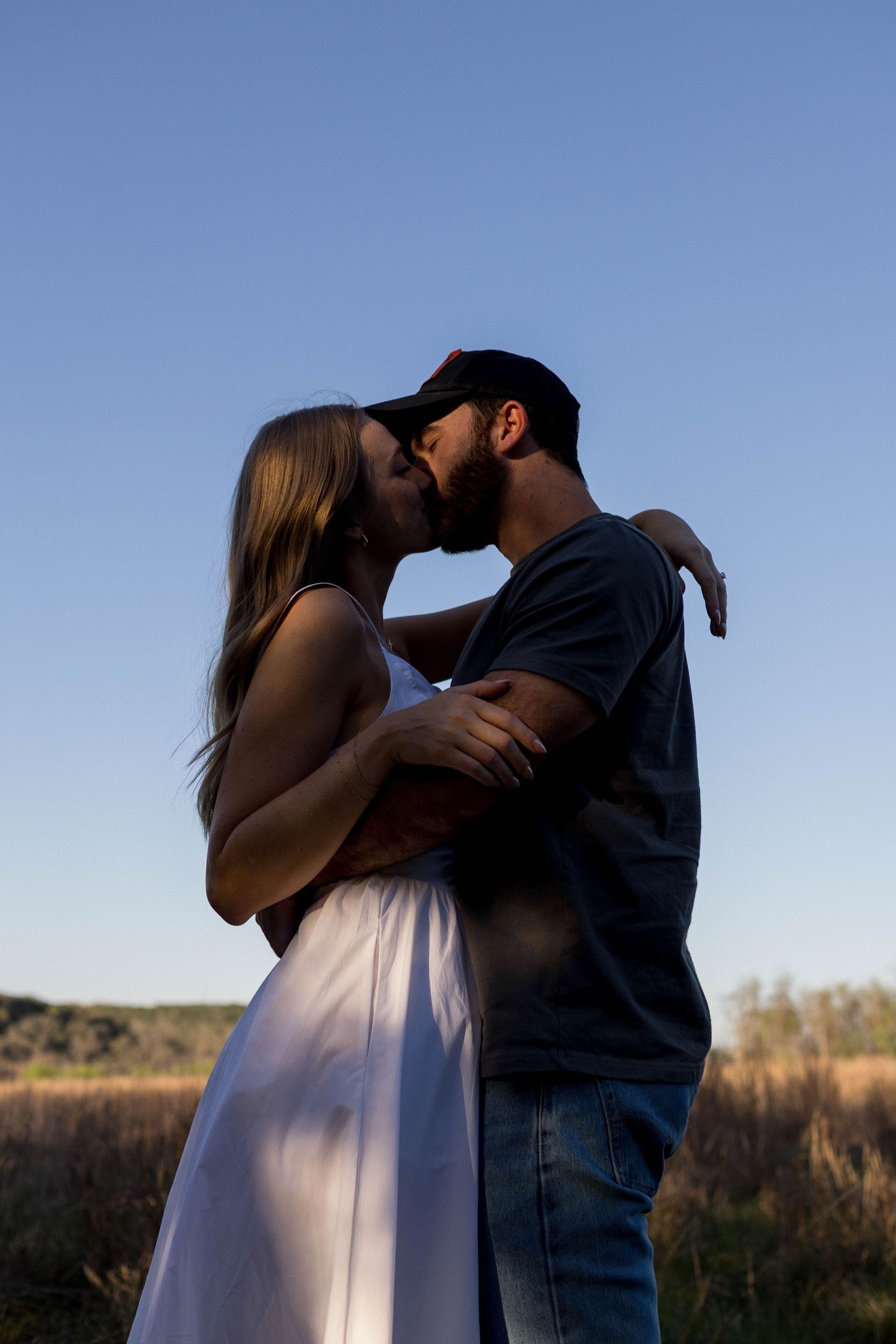 Couple sharing a romantic kiss outdoors in a field at sunset, woman in white dress, man in cap and jeans.
