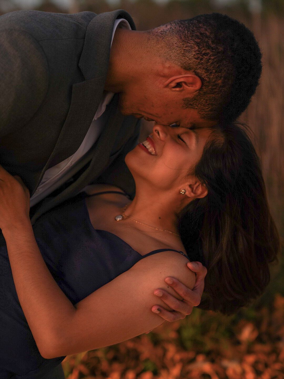A couple shares a romantic embrace outdoors, the man leaning in to kiss the smiling woman in warm golden light.