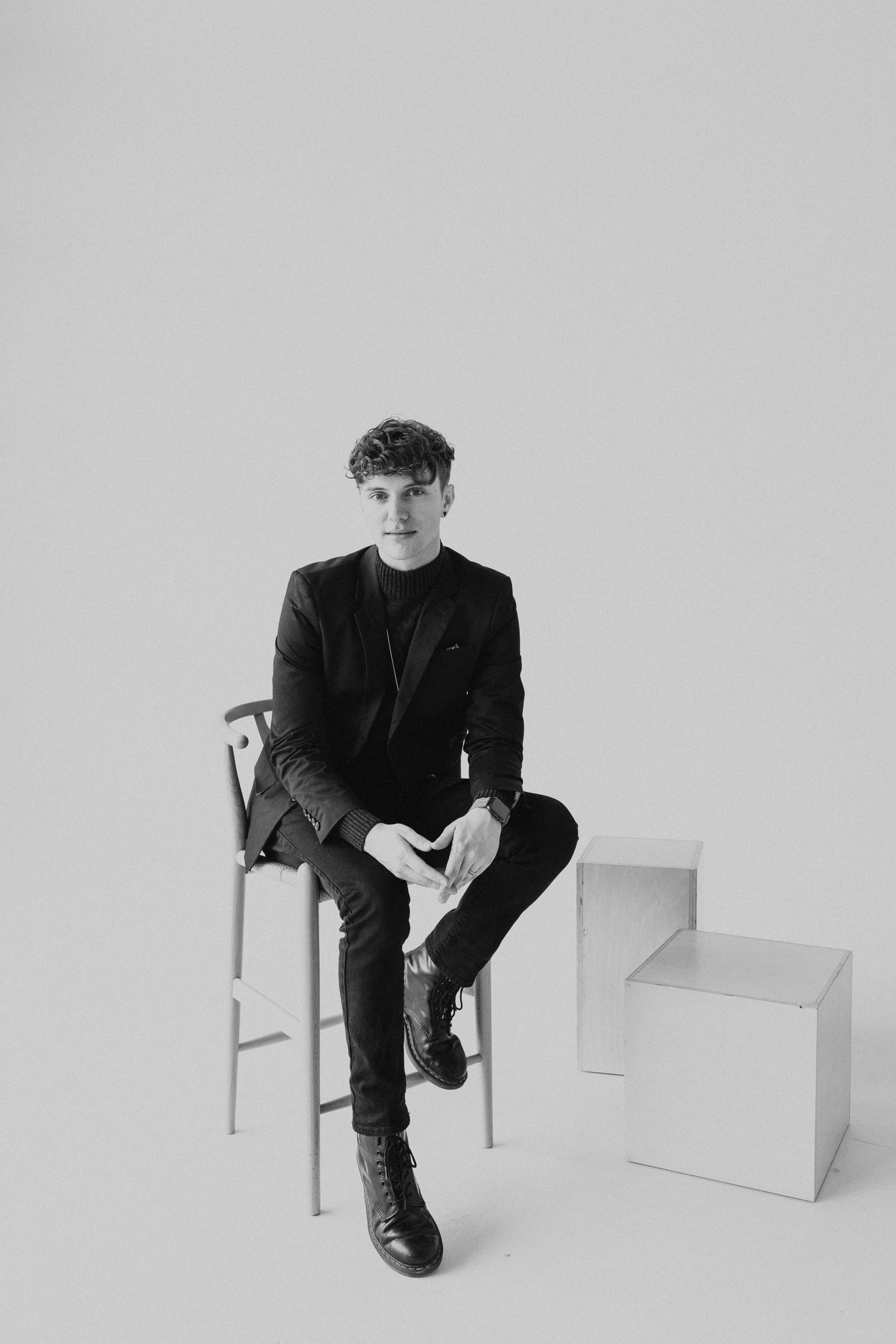 Young man in black suit sitting on a bar stool in a minimalist black and white studio portrait.