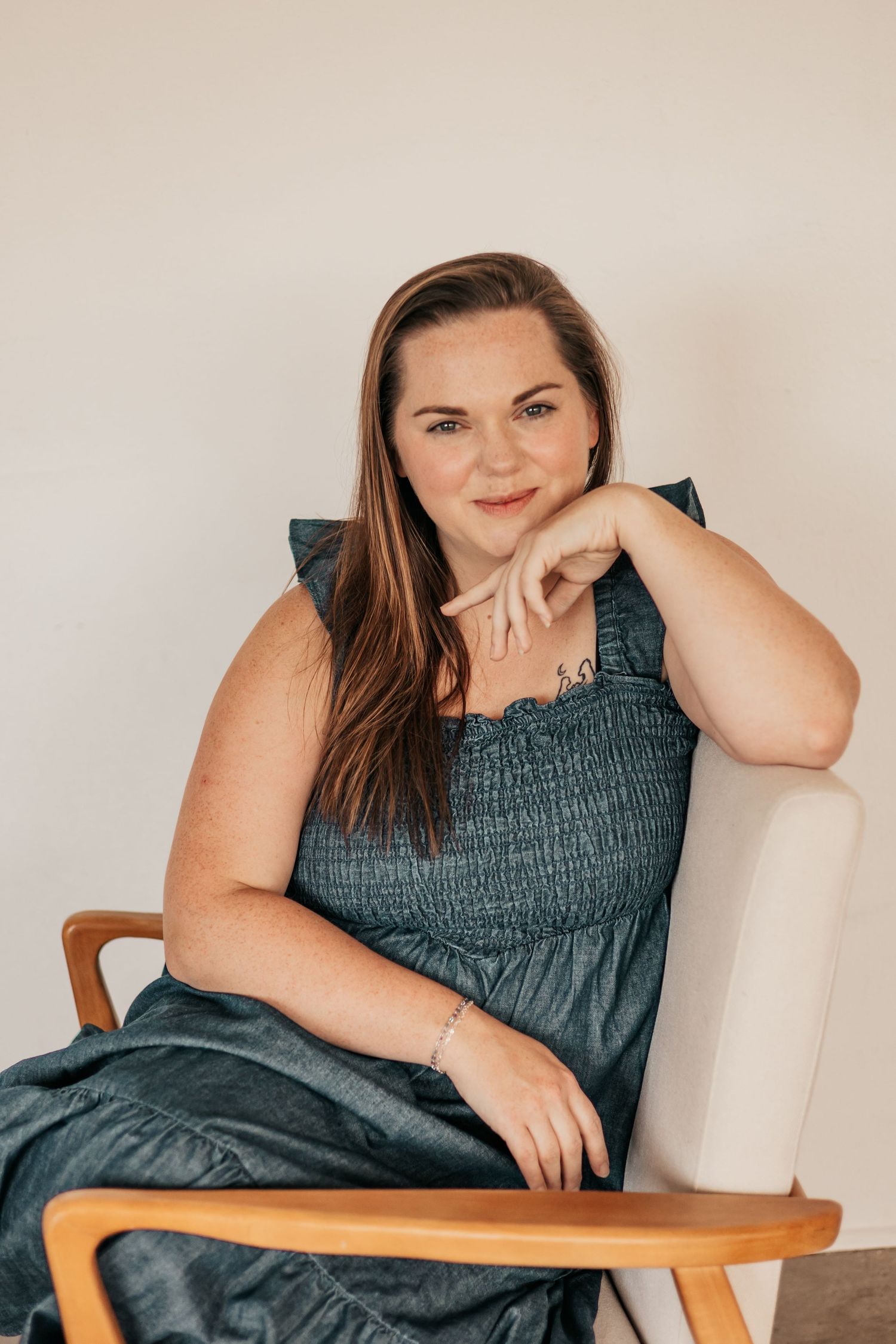A smiling woman in a teal smocked dress sits relaxed in a wooden chair against a light background | Photography Business Coach