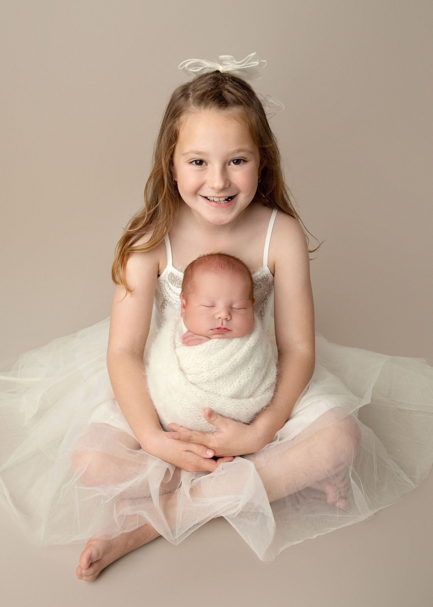 Young girl in white tutu dress holds swaddled sleeping newborn baby sibling in a professional studio portrait.