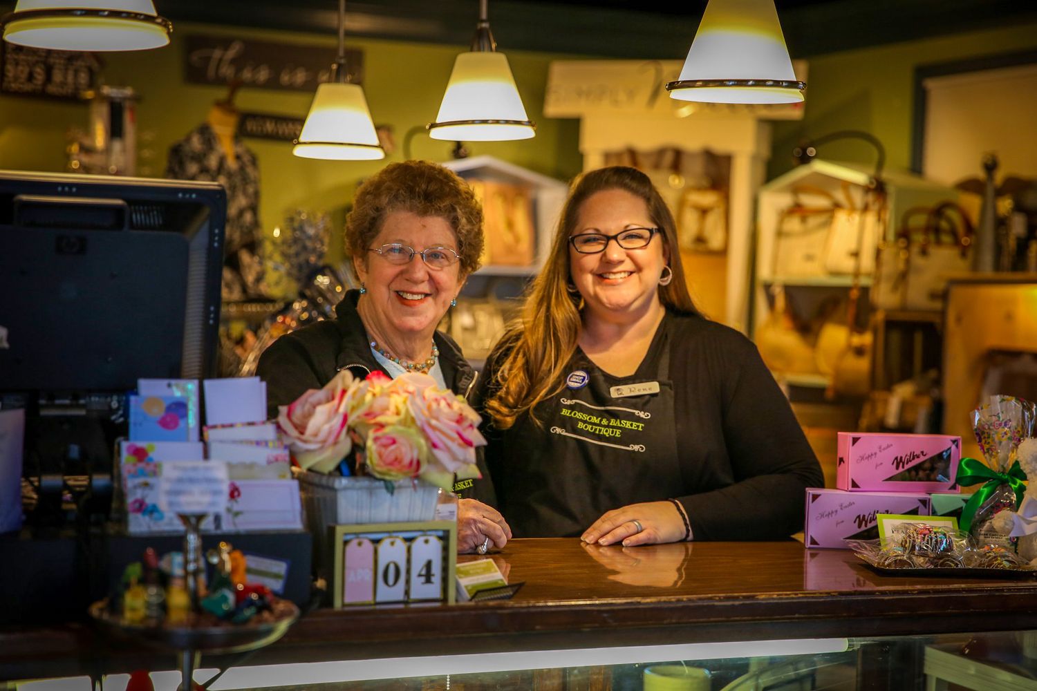 Two smiling women pose together behind a retail shop counter surrounded by colorful gifts and merchandise.