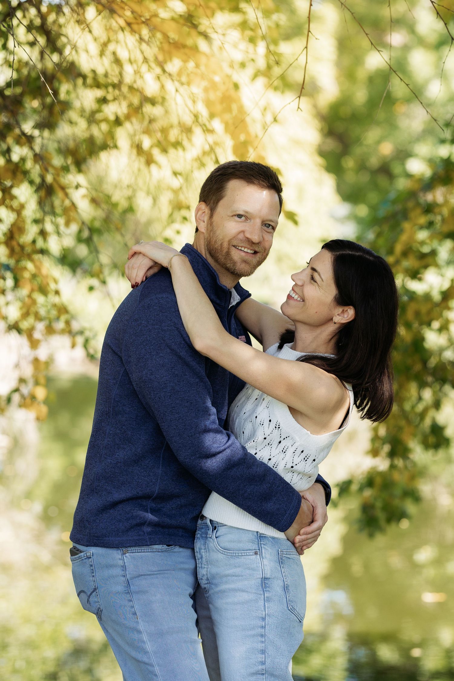 A smiling couple embraces outdoors among lush green trees, wearing casual jeans and tops in a park setting.