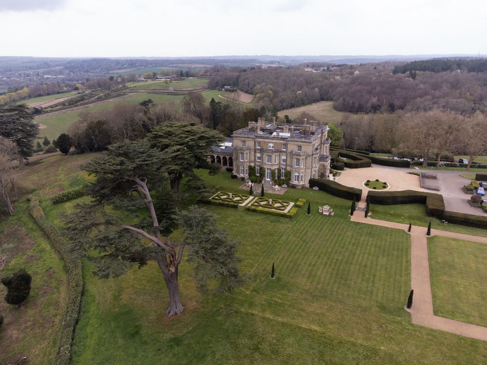 Aerial view of a grand English country manor house surrounded by manicured gardens and rolling countryside.