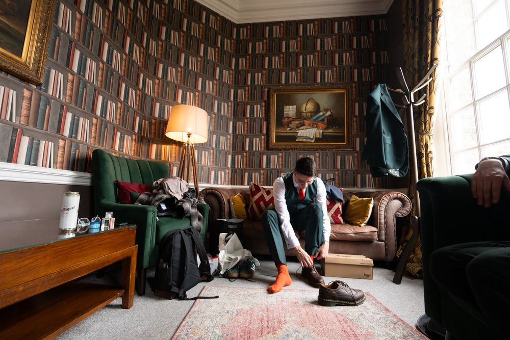 Groom getting dressed in a book-wallpapered room, sitting on a green sofa tying his shoes before the wedding.