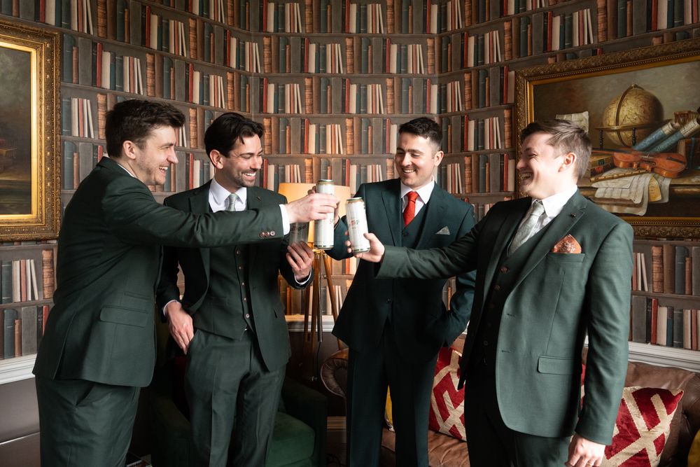 Four groomsmen in dark green suits toasting with champagne in an elegant library setting.
