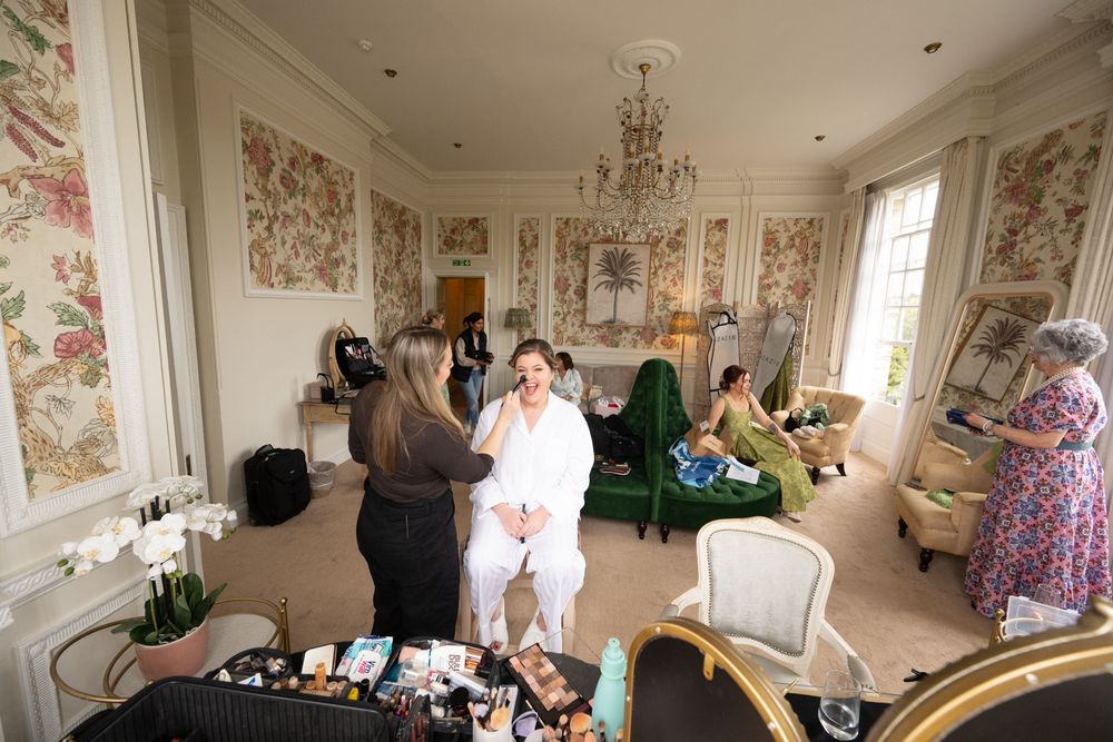 Bride getting makeup done in an ornate room with floral wallpaper and chandelier surrounded by wedding party members.