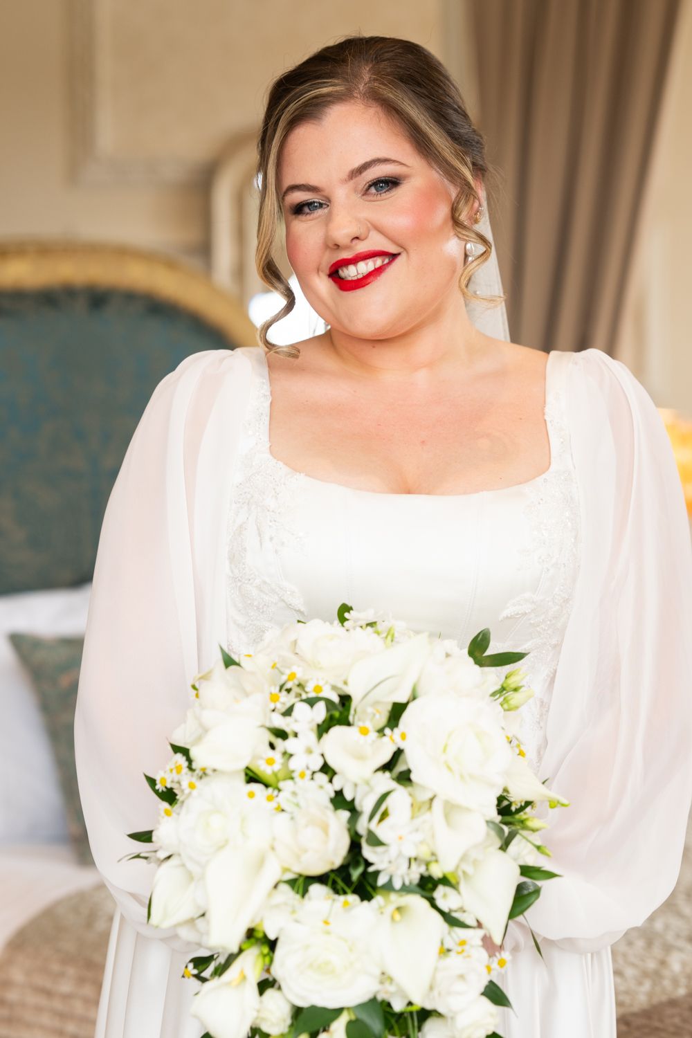 Smiling bride in white gown with red lipstick holding a white rose bouquet indoors.