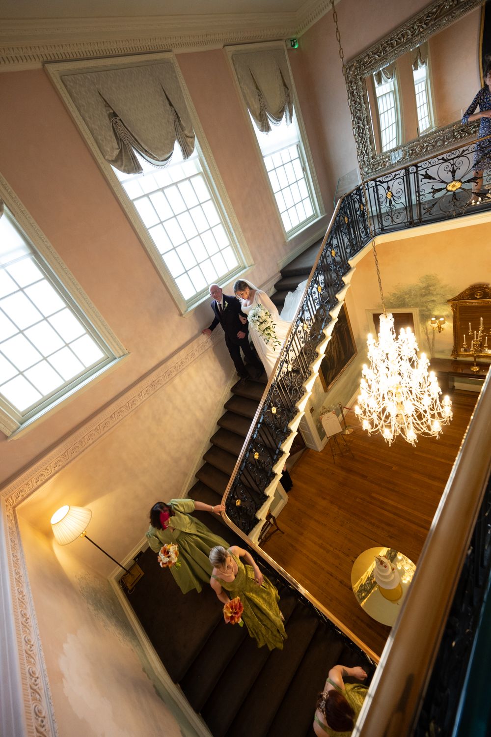 Aerial view of elegantly dressed wedding party descending grand staircase with chandelier in historic venue.