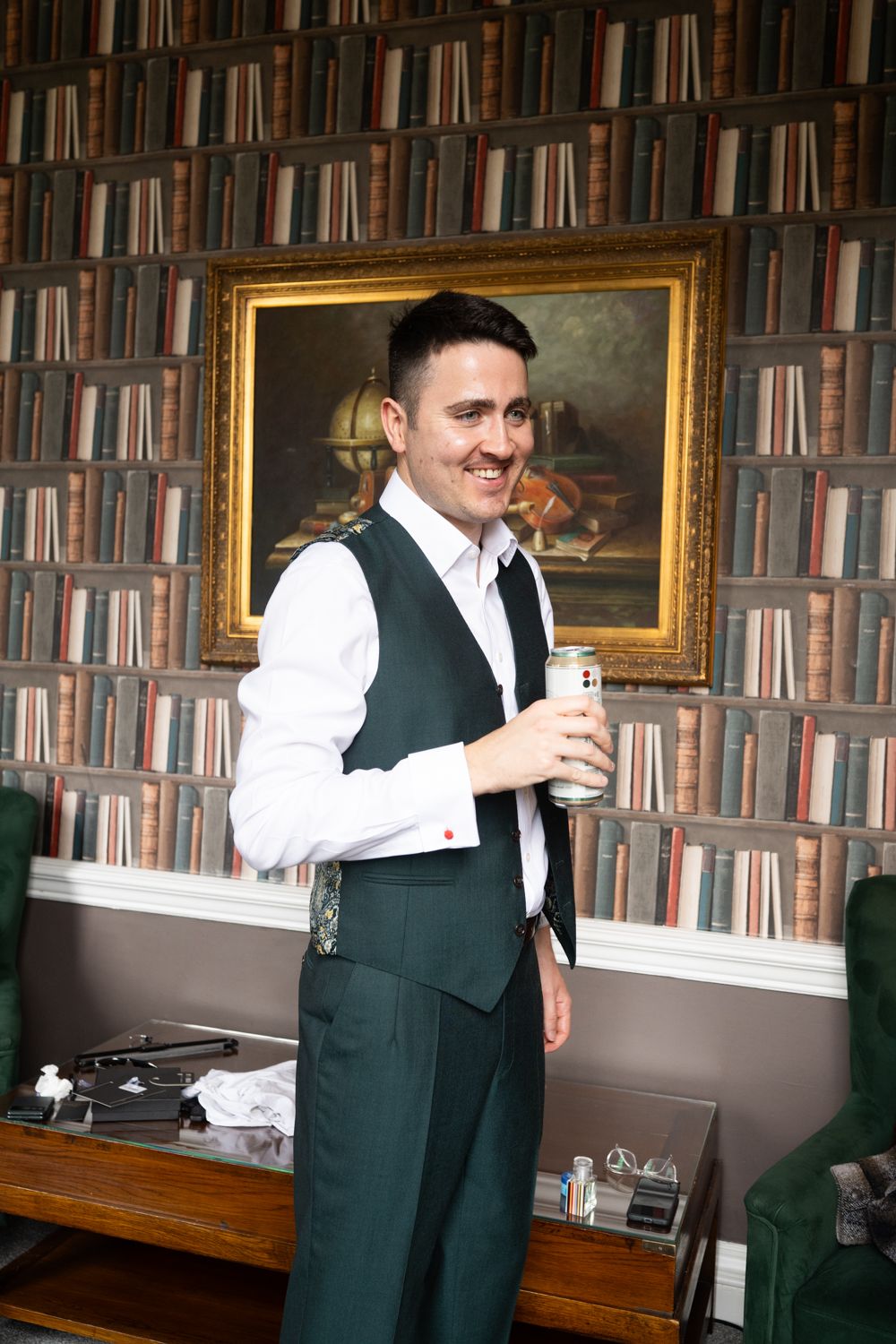 A smiling man in a green vest and white shirt holds a drink in a library-themed room with a gold-framed painting.