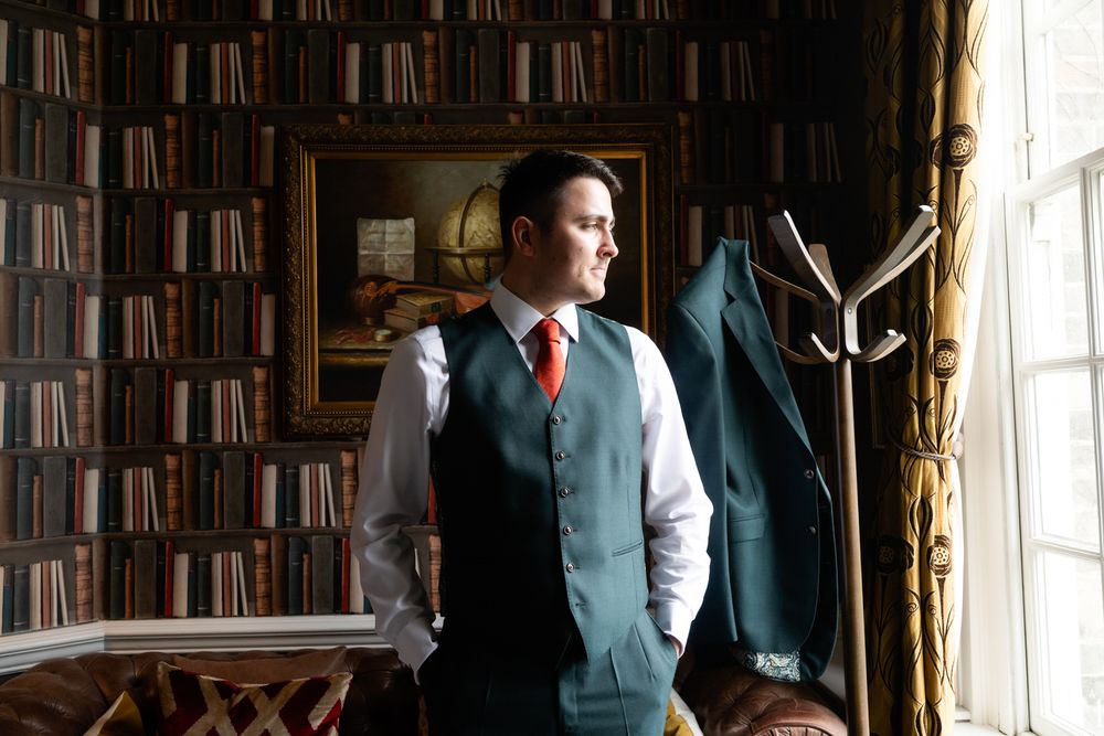 Groom in green vest and red tie stands by bookshelf window, suit jacket on coat rack nearby.