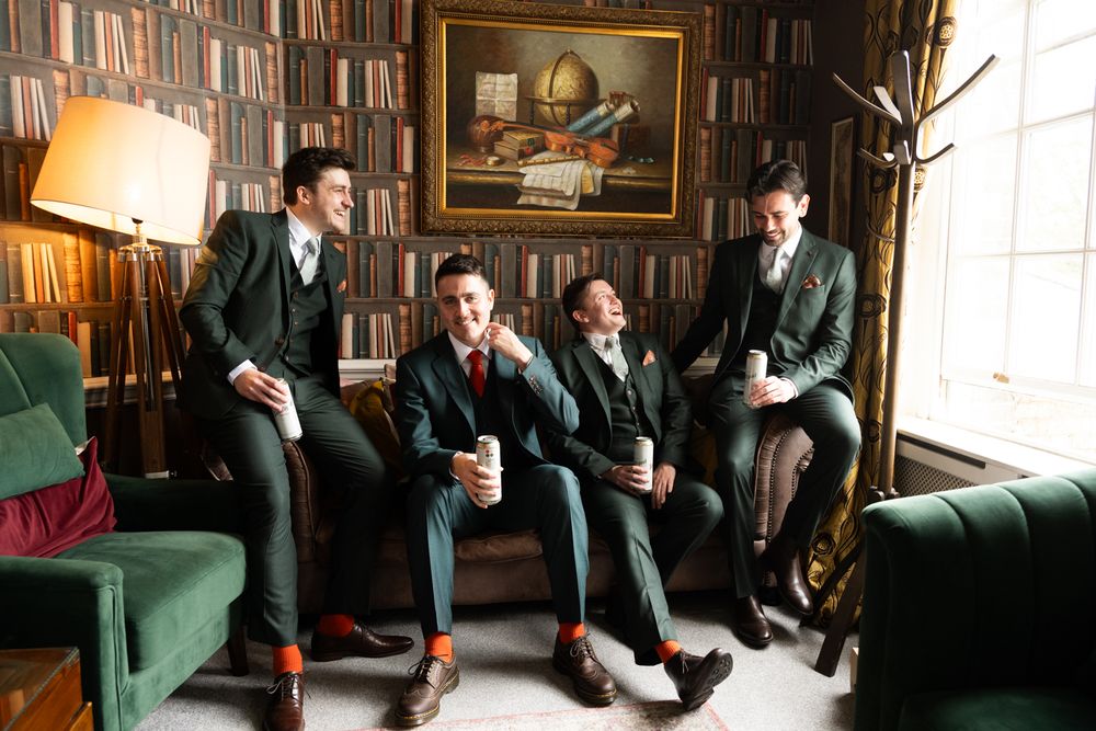 Four groomsmen in dark suits with red ties laugh together on a green sofa in a library setting.