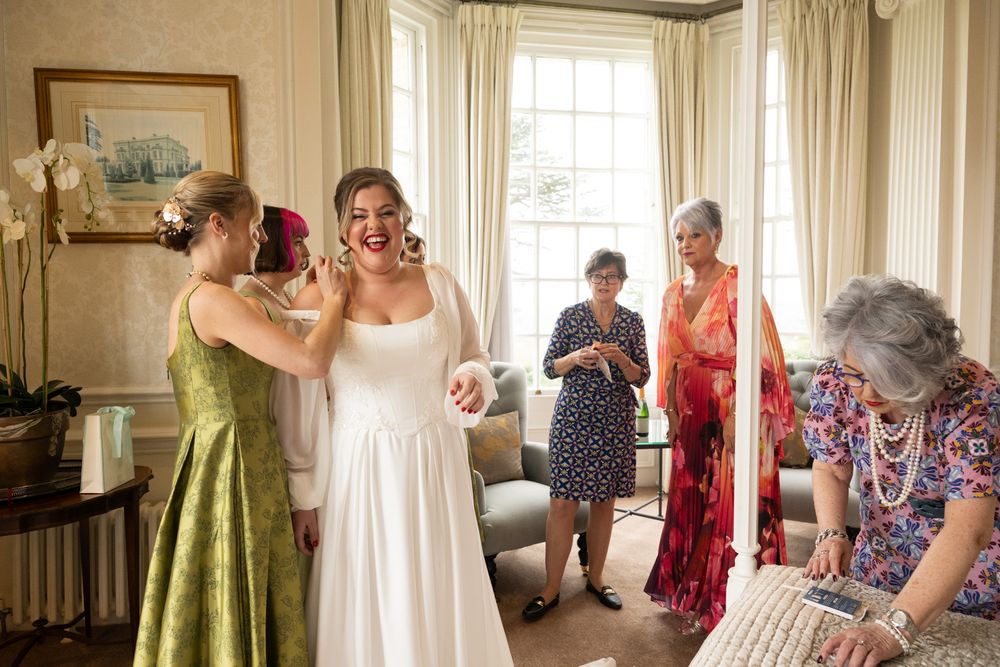 Bride laughing joyfully as bridesmaids help her dress, surrounded by family in an elegant room.