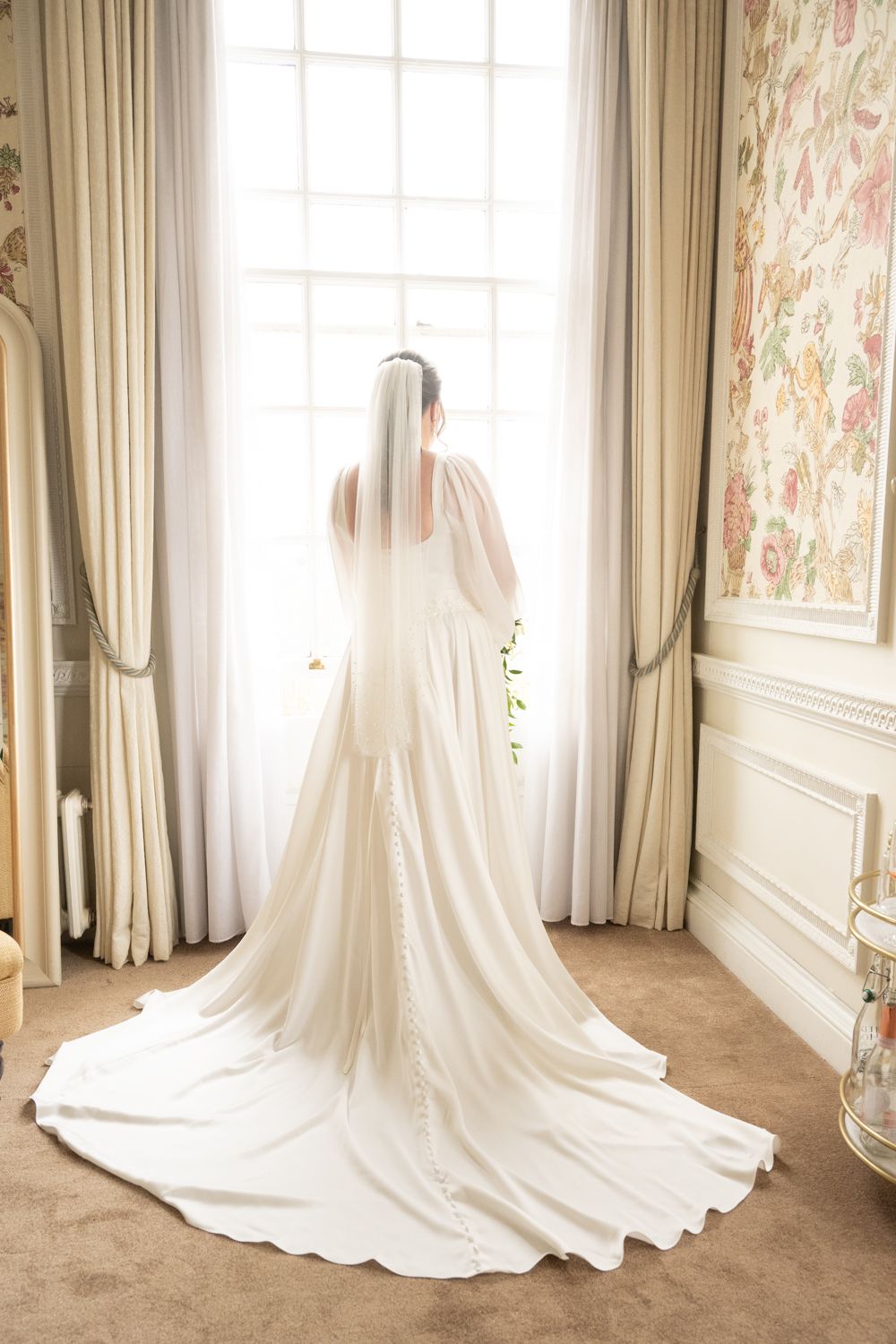 Bride in elegant white wedding gown with long train and veil stands before a bright window in a floral-wallpapered room.