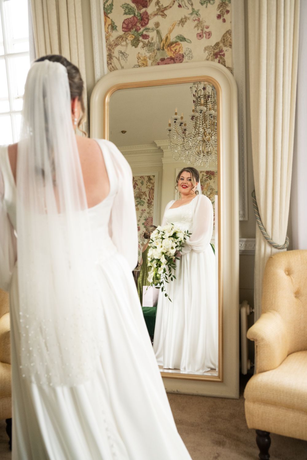 Bride in white gown and veil holds bouquet, smiling at her reflection in an ornate gold-framed mirror.
