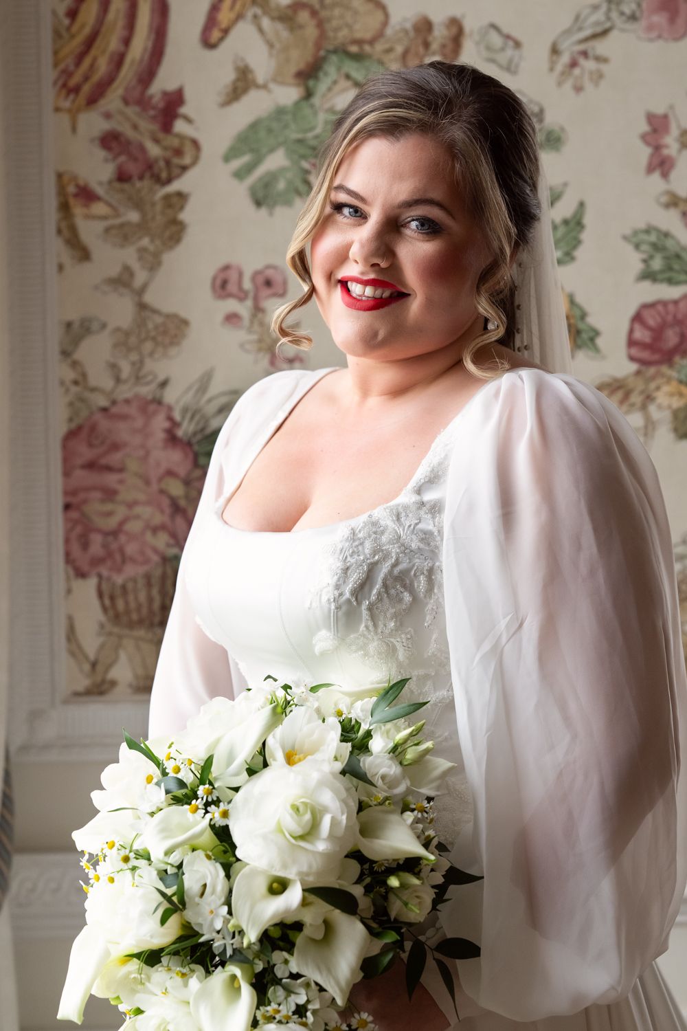 Smiling bride in white gown holding bouquet of white roses and calla lilies, standing before floral wallpaper.