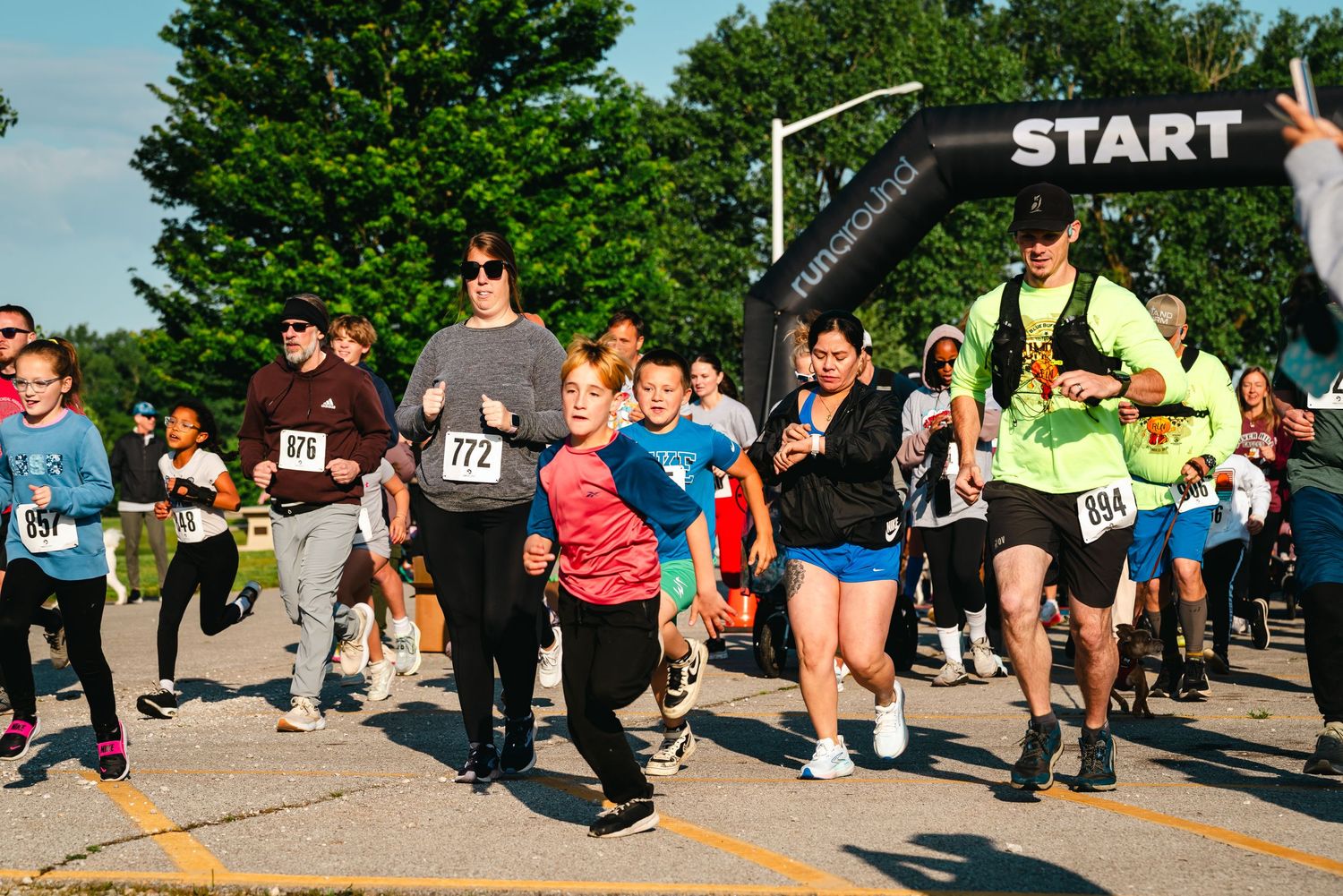 Runners crossing the start line at an outdoor 5K race event on a sunny day.