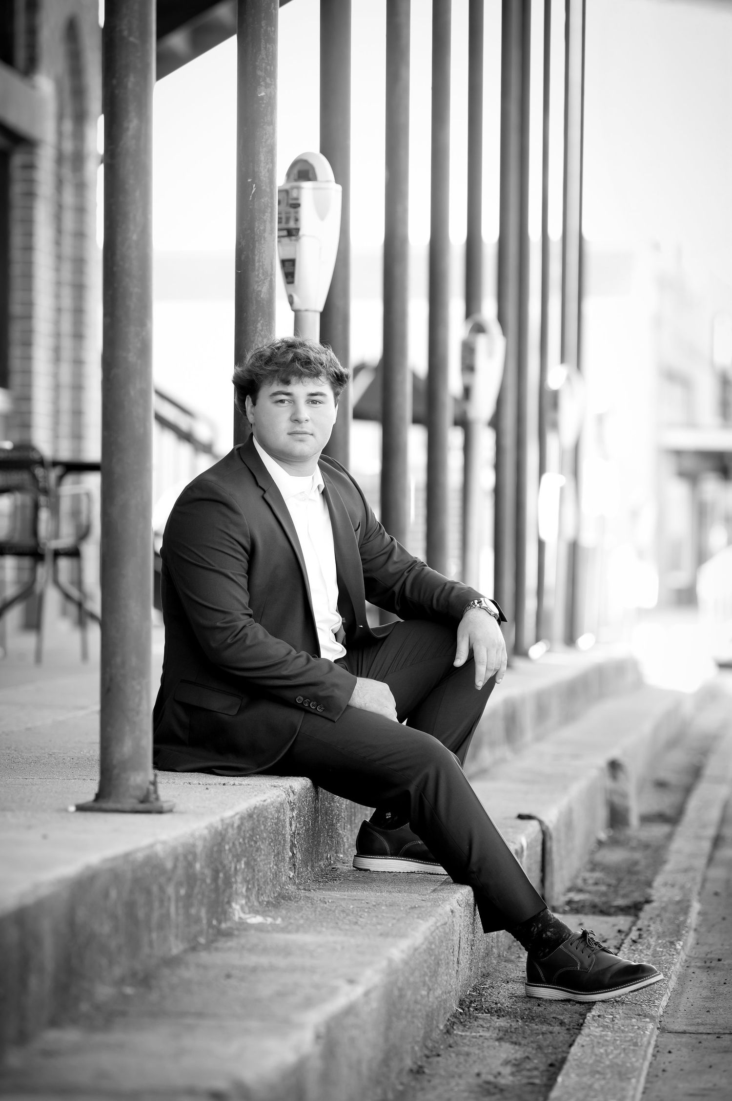 Young man in black suit sitting on steps in black and white senior portrait photo.