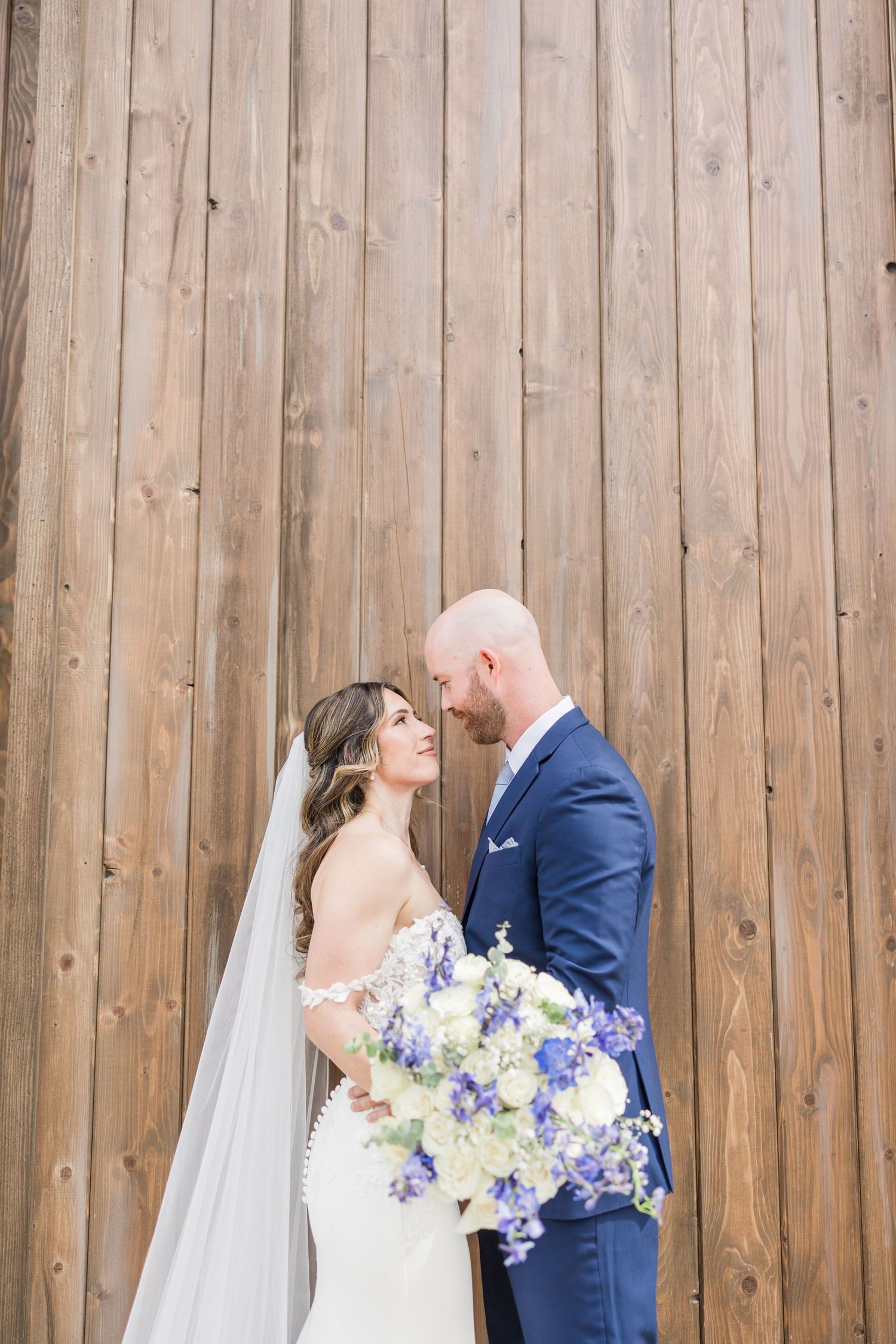 Bride and groom share a romantic moment against a rustic wood wall, bride holding blue and white floral bouquet.