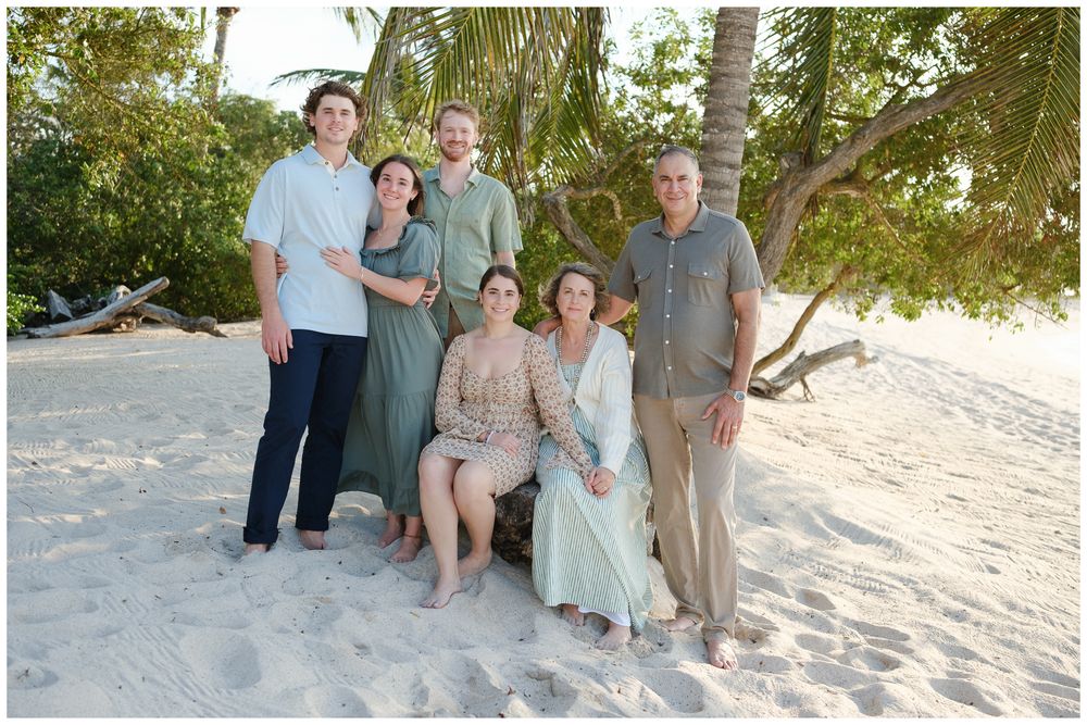 Family of six posing together on a sandy tropical beach with palm trees, wearing coordinated earth-tone outfits.
