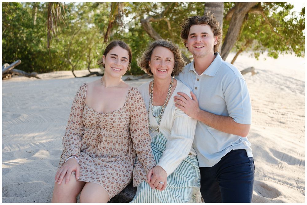 A family of three smiles together on a sandy beach, with two young adults flanking an older woman in soft, natural tones.