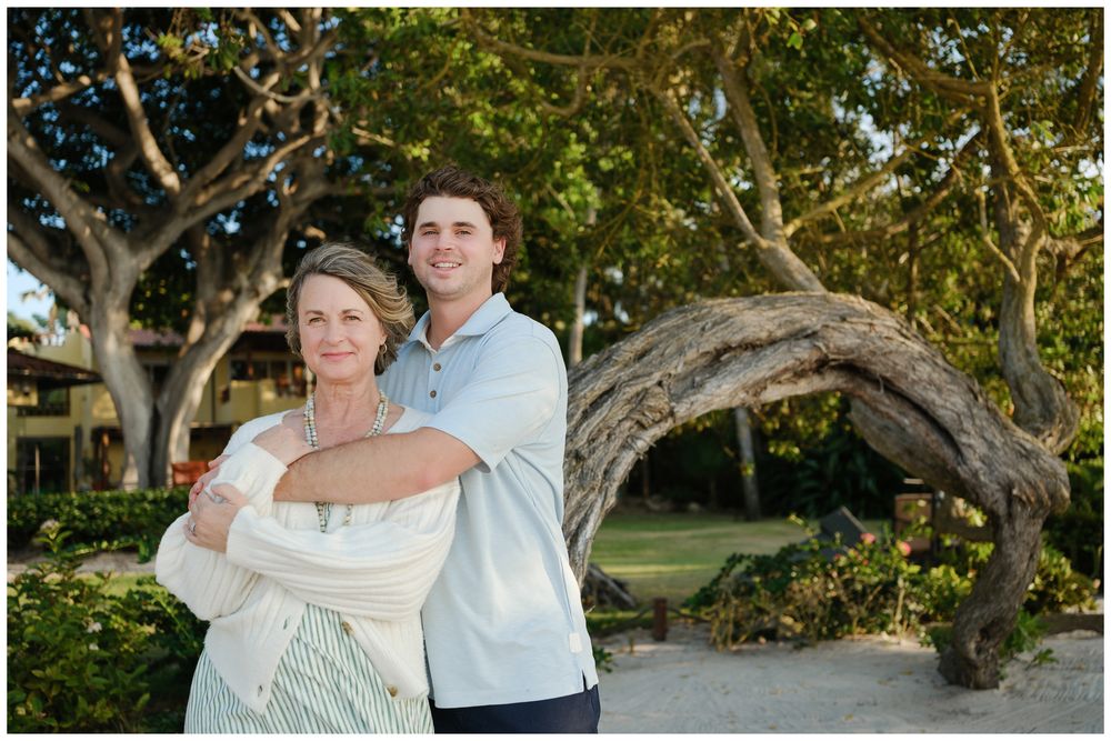 An older woman and young man pose together outdoors near a scenic twisted tree in a tropical garden setting.