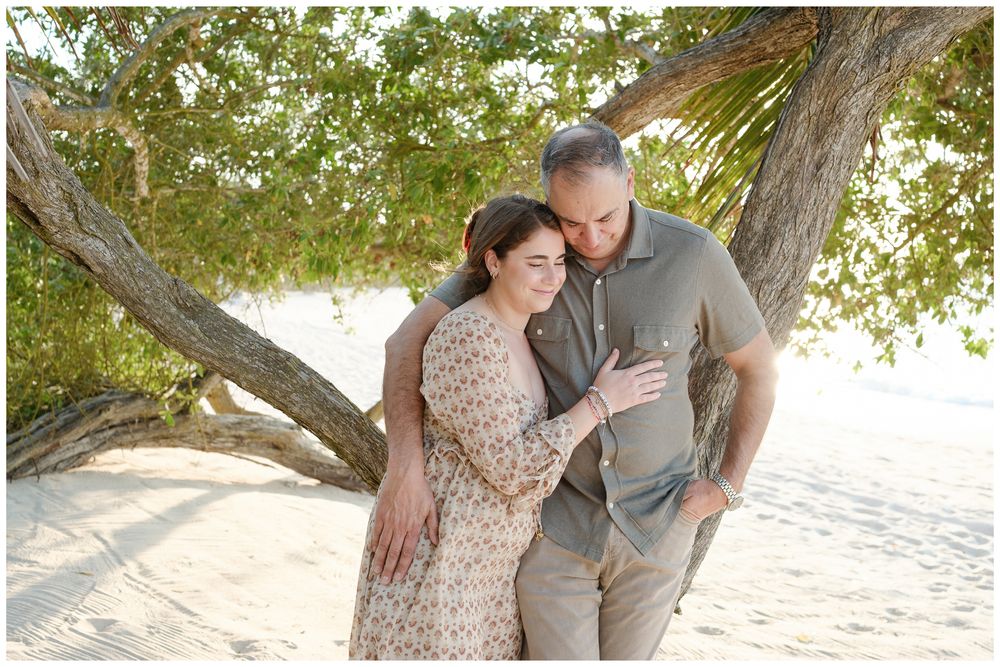 Couple embracing under a tropical tree on a white sandy beach, sharing a tender romantic moment.