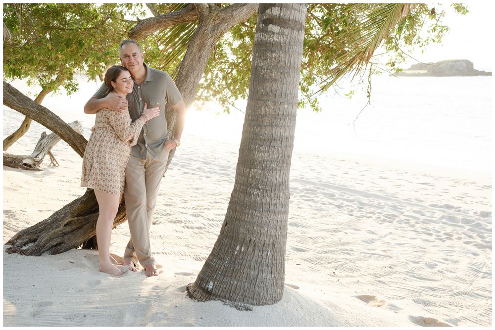 Couple embracing by a leaning palm tree on a white sandy tropical beach during golden hour.