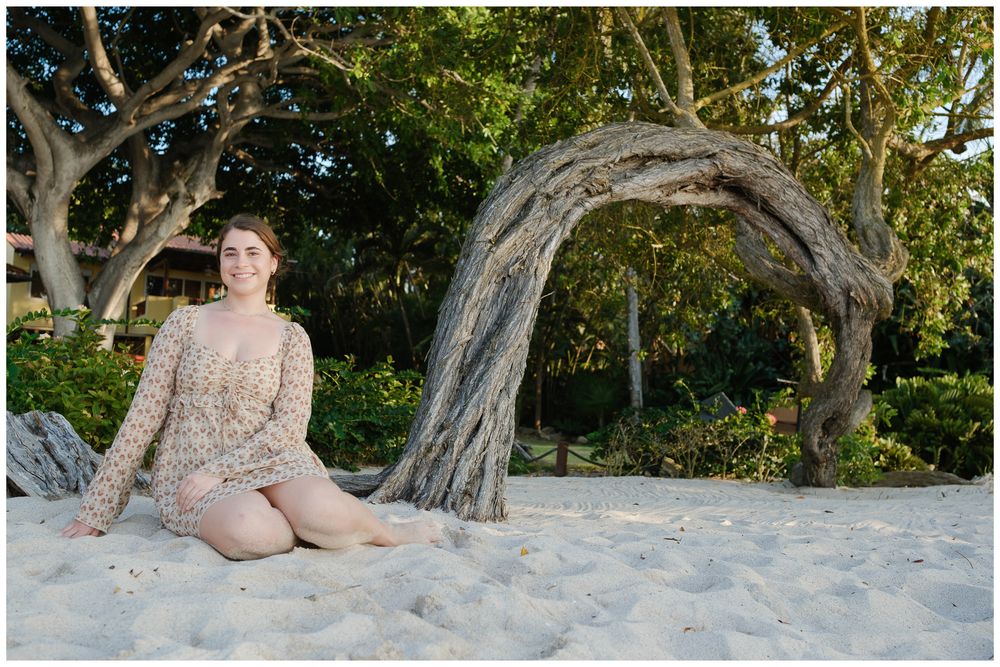 Young woman in floral outfit sitting on white sand beach with arched driftwood tree and lush greenery backdrop.