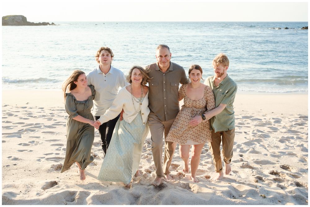Happy family of six posing together on a sandy beach at sunset, smiling and embracing in casual summer attire.