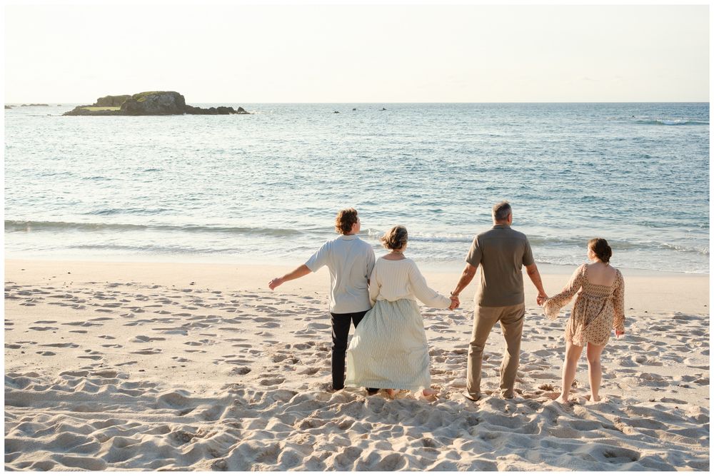 Four people holding hands walk toward the ocean on a white sandy beach at sunset.