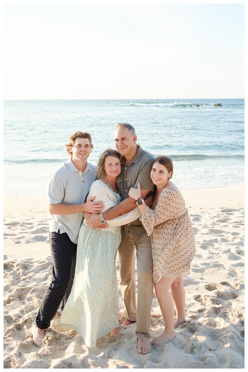 Family of four smiling together on a sandy beach with ocean waves in the background during golden hour.