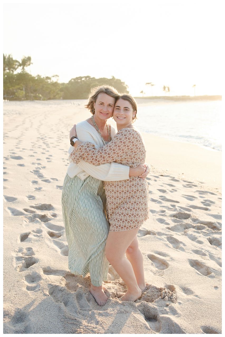 Two women hugging and smiling on a sandy beach at golden hour, wearing casual summer dresses.