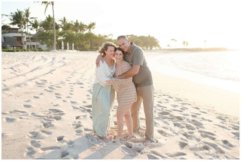 Family of three embracing and smiling on a white sandy beach at golden hour with palm trees in background.