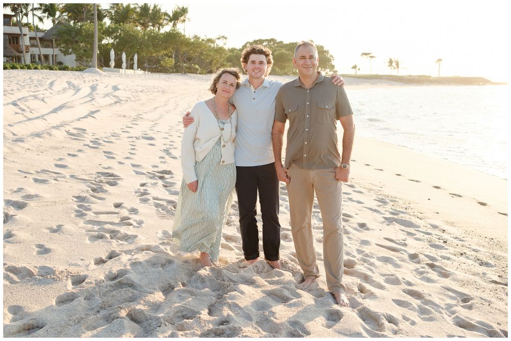 Three people smiling together on a sandy tropical beach with palm trees and a resort in the background.