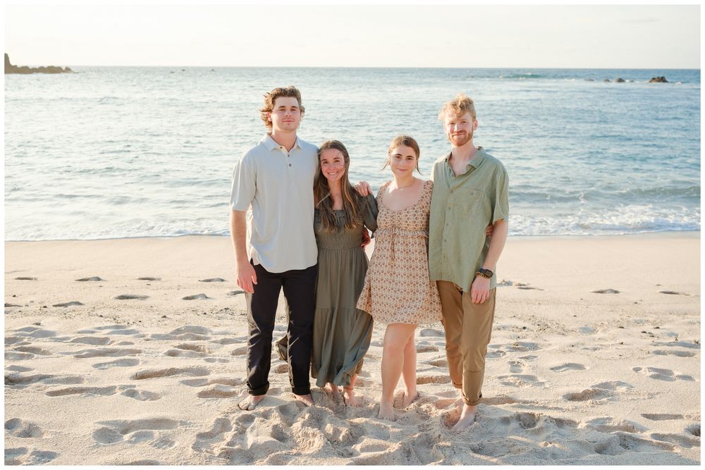 Four people posing together on a sandy beach with calm ocean waves and a pastel sky in the background.
