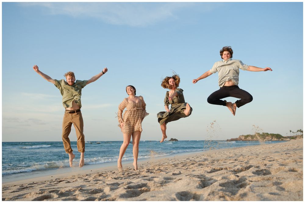 Four joyful friends jumping in the air on a sunny beach with blue skies and tropical scenery.