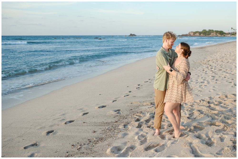 Couple embracing on a sandy beach at sunset with gentle waves and footprints in the sand behind them.