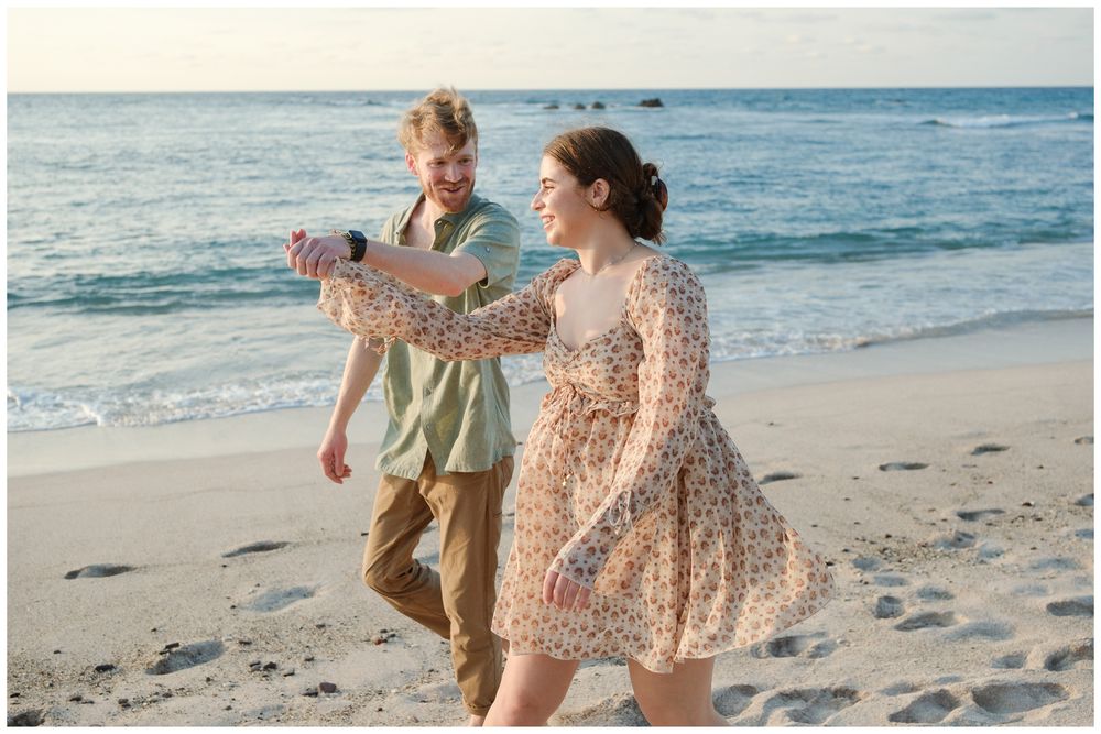 Couple walking on a sandy beach, man holding a camera, woman in a floral dress, ocean waves in the background.