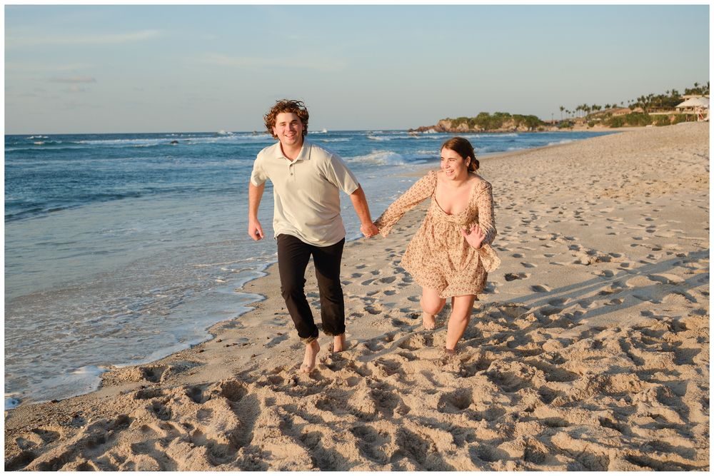 Couple holding hands and running joyfully along a sandy beach at sunset with ocean waves in the background.