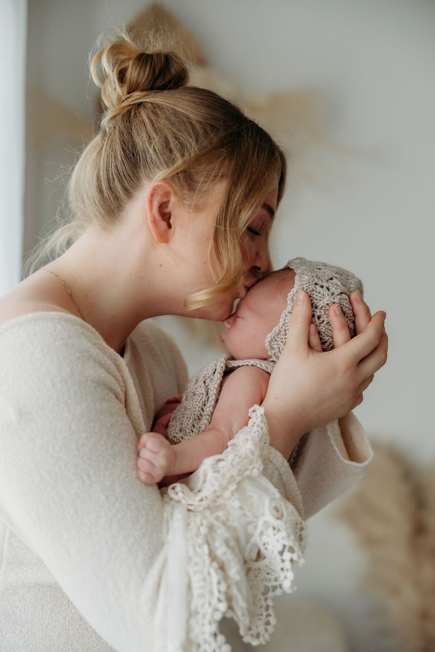 A tender moment as a young blonde mother kisses her newborn baby wrapped in a delicate lace knit blanket.