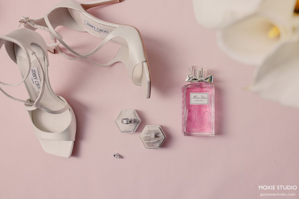Bridal flat lay with white heels, diamond rings, hexagon ring boxes, and pink Miss Dior perfume on a pink background.