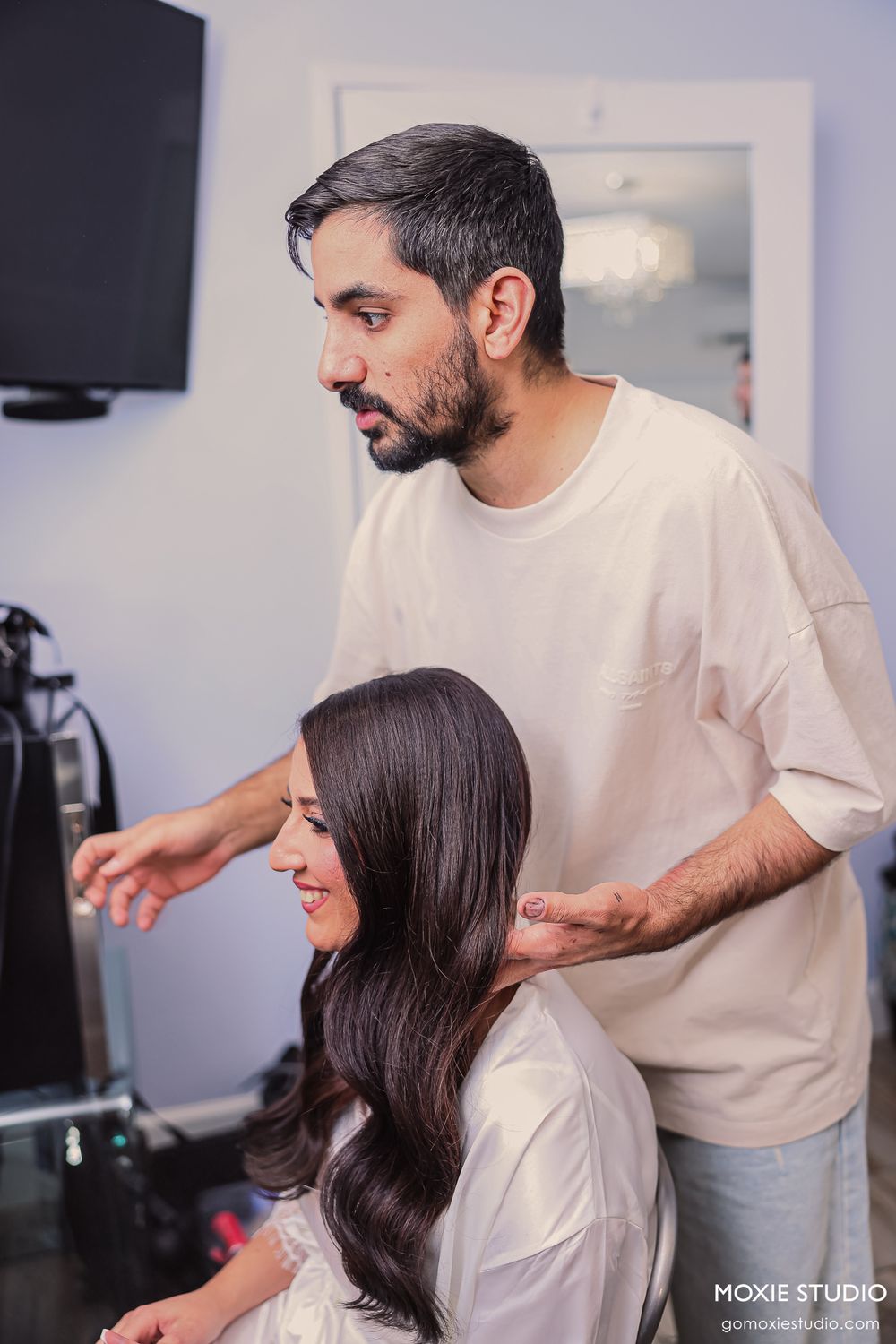 A hairstylist styling a woman's long dark hair in a bright salon setting.